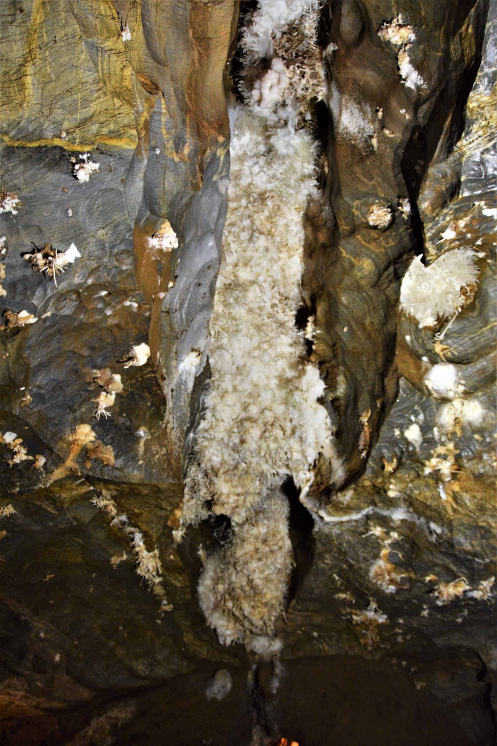 massive white Aragonite structure growing inside a crevice in the cave ceiling resembling mould surrounded by blue marble and smaller Aragonite clusters growing out of holes