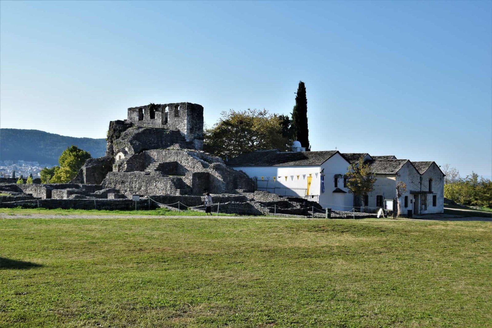 a green yard in front of the remains of a medieval tower next to a restored building complex with black slate-tiled roofs