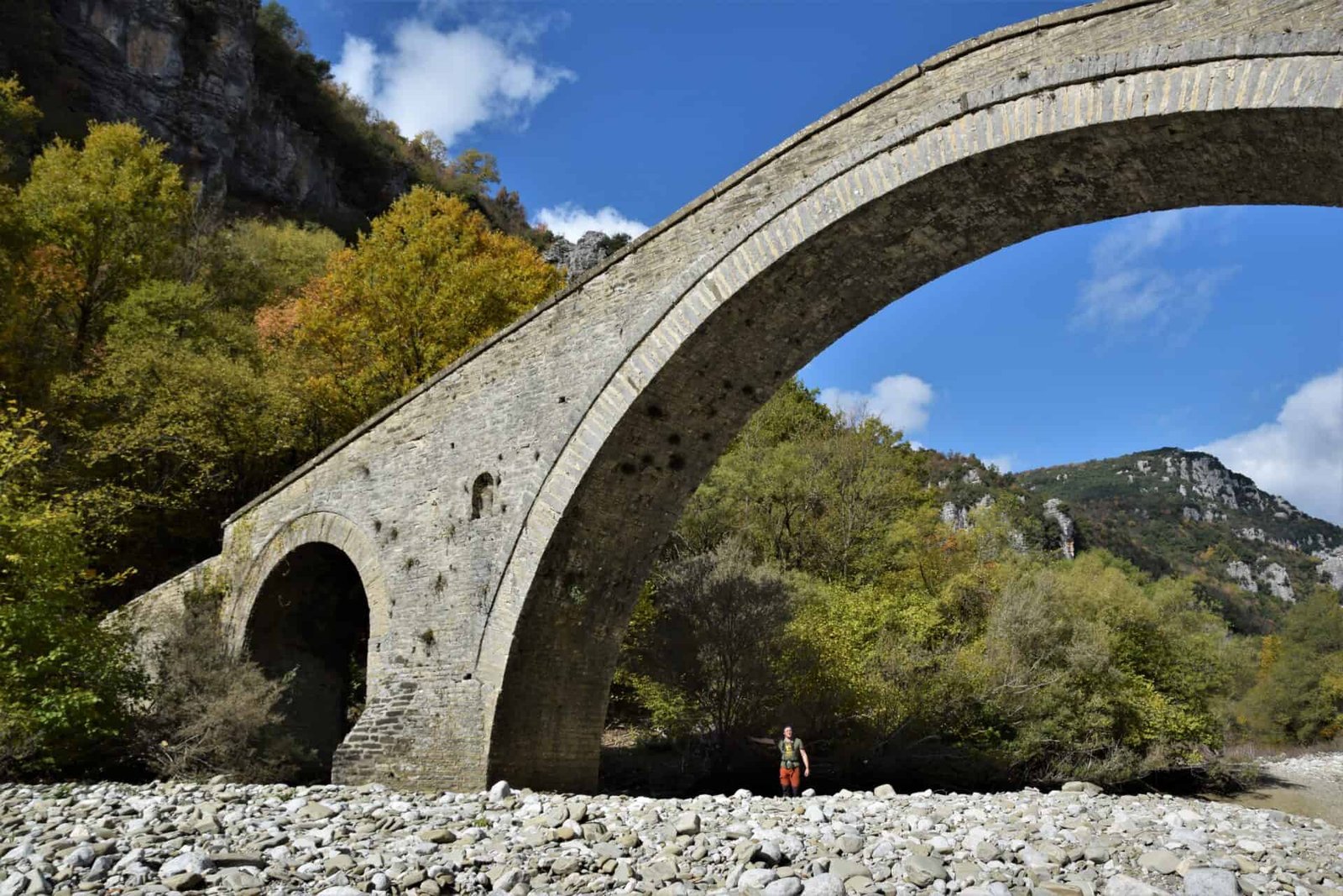 a hiker, wearing red trousers and a green shirt, stands beneath a large stone bridge arching across a dry riverbed in a canyon