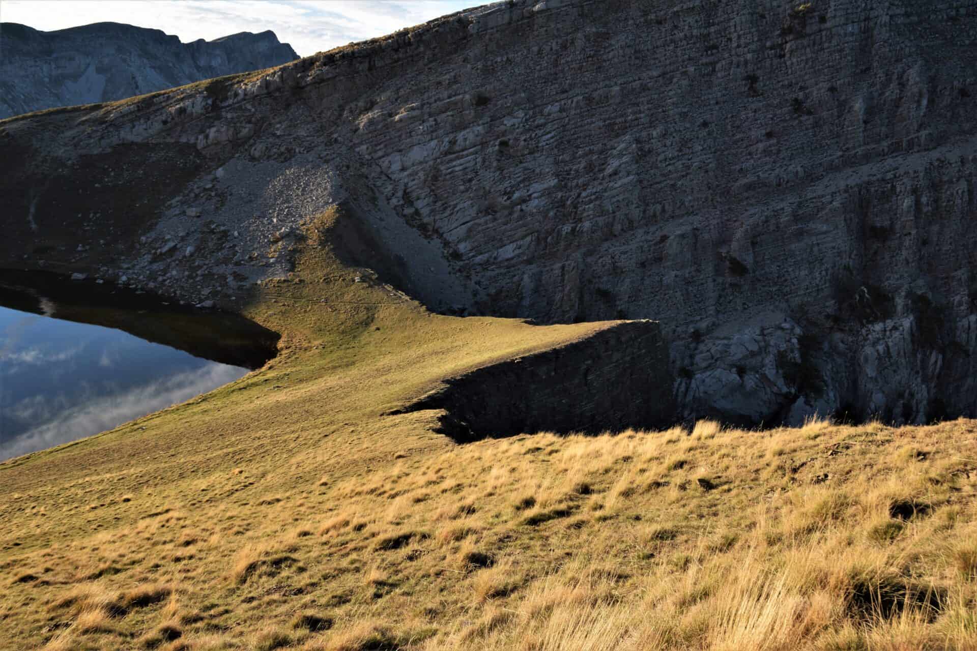 pointy terrain leading up to a vertical drop in front of another steep cliffside next to an alpine lake