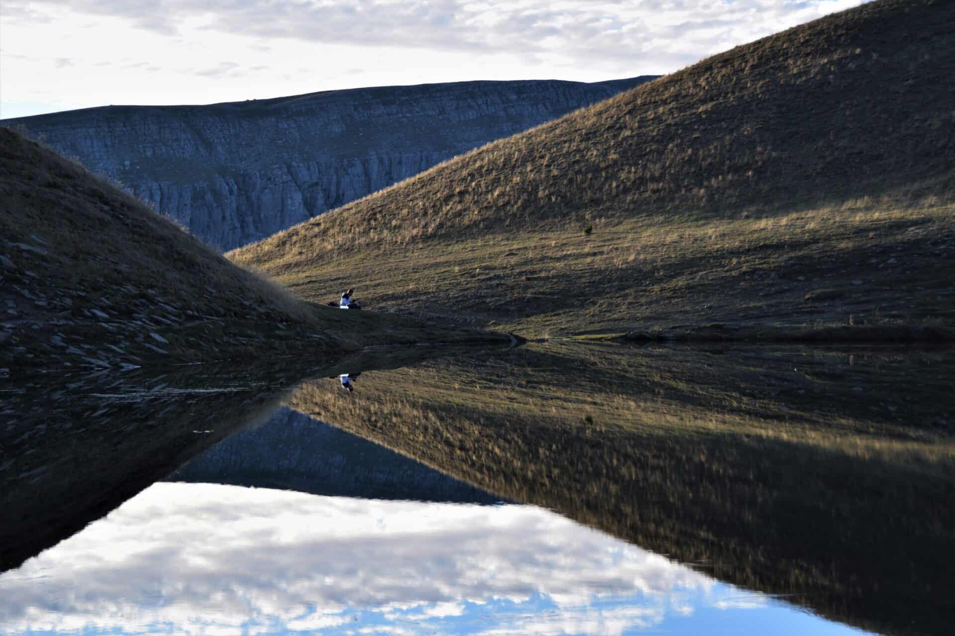 man in a white jacket, surrounded by brown meadows, sits right next to a mountain lake, mirroring the scenery