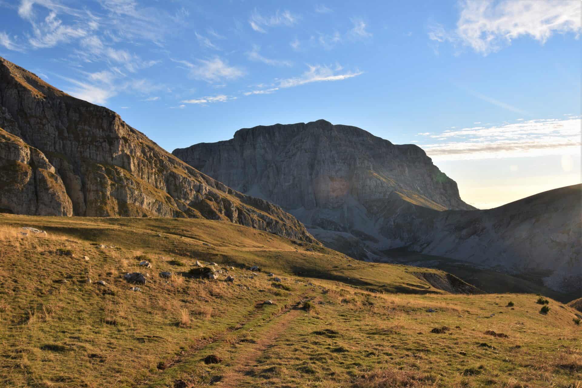 dirt path leads down mountain pastures bathed in warm light towards a enormous mountain massif