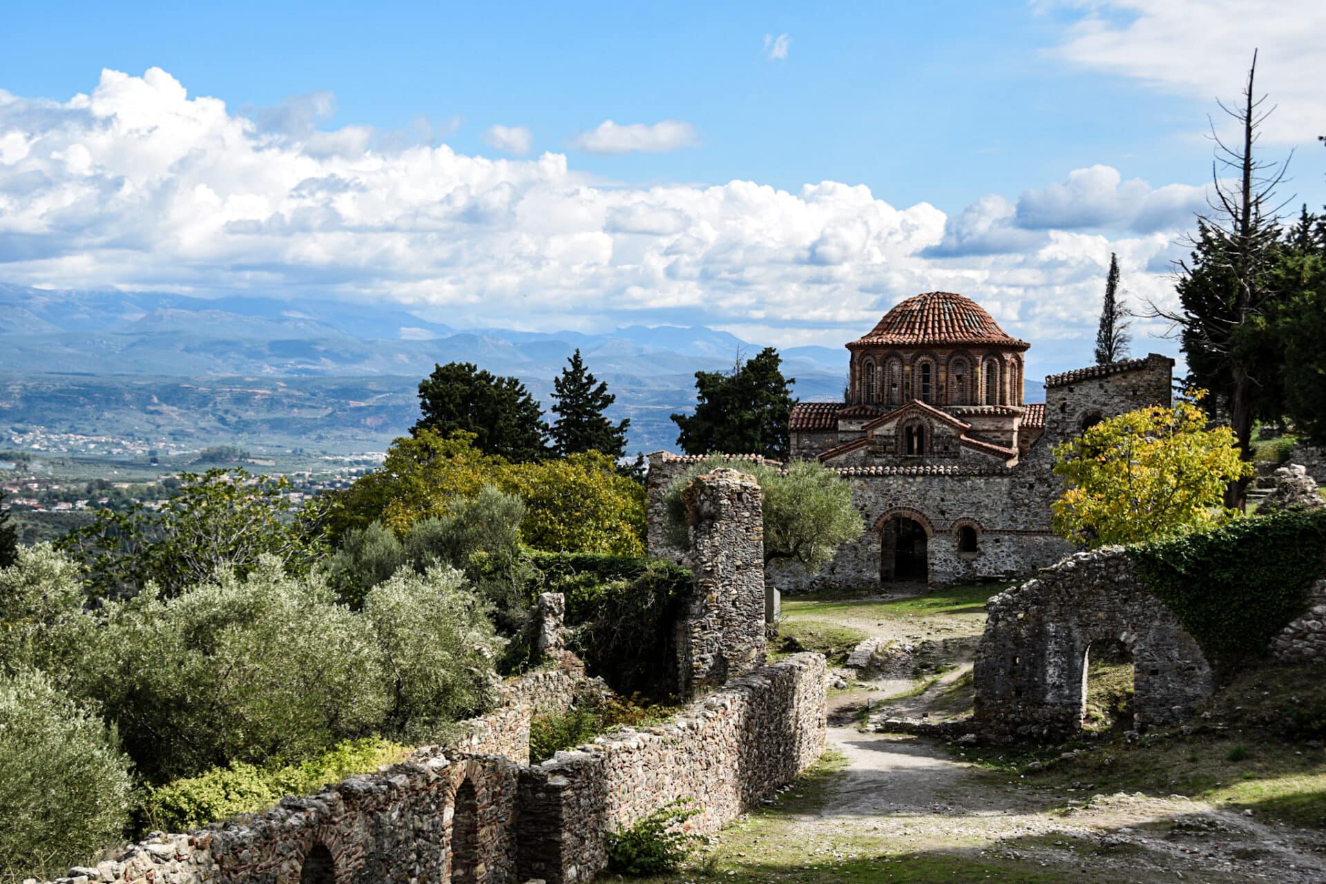 Byzantine monastery boasting a red tiled dome and roofs behind a collapsed wall surrounded by lush trees