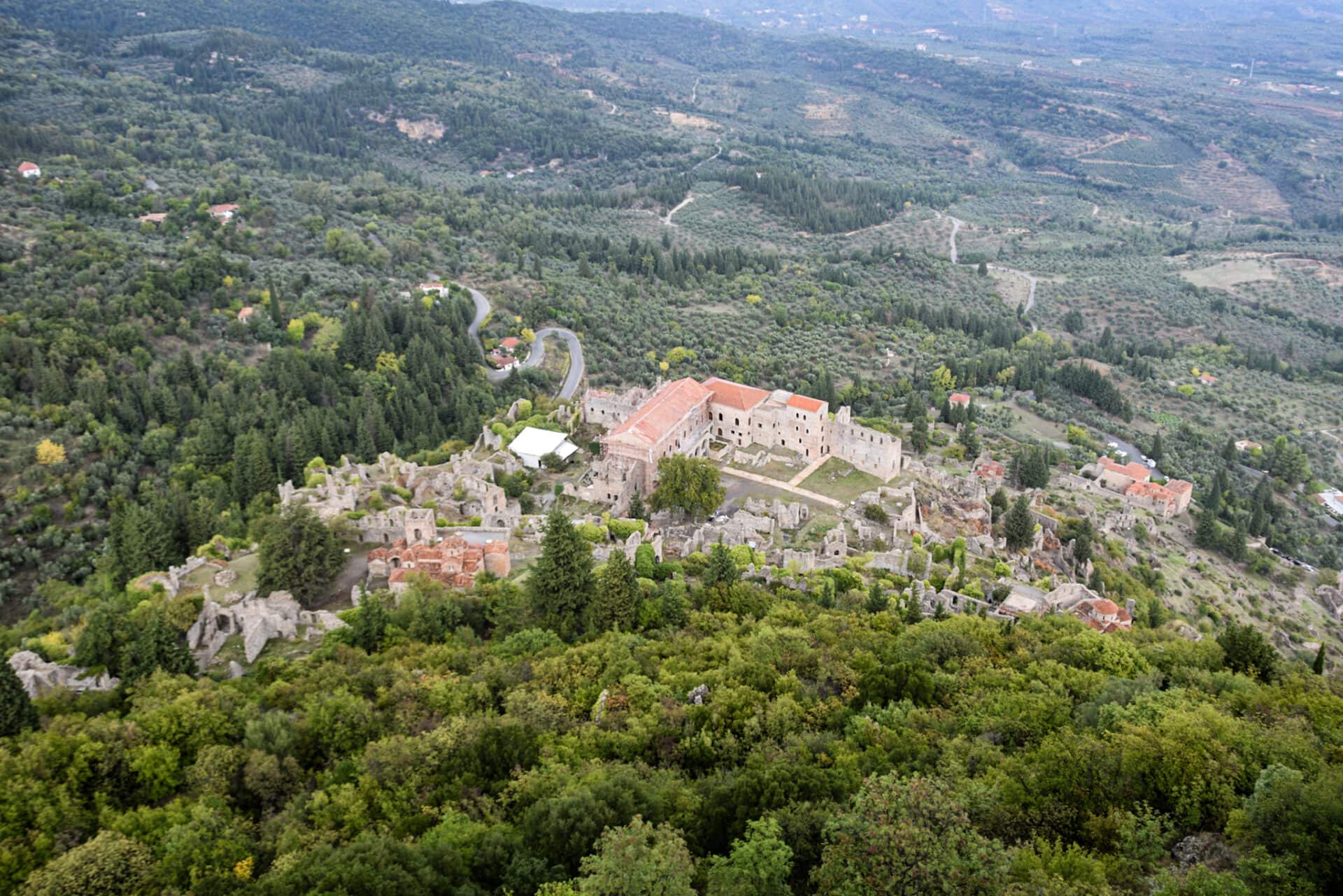 a palace and several other ruined buildings hug the forested slopes of a hill overlooking a fertile plain covered in olive groves