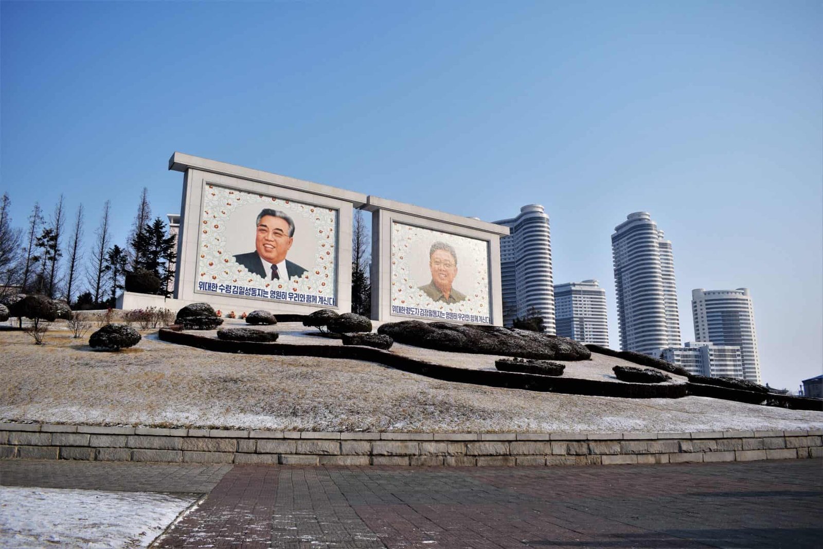 two giant portraits of former North Korean leaders Kim Il-sung and Kim Jong-il behind an ensemble of boxtrees and in front of blue coloured skyscrapers