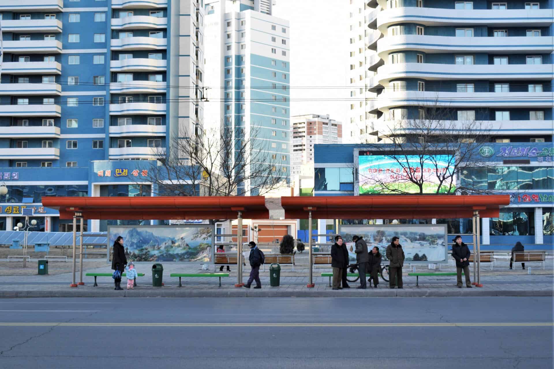 people waiting for the bus in front of blue coloured buildings in North Korea's capital Pyongyang