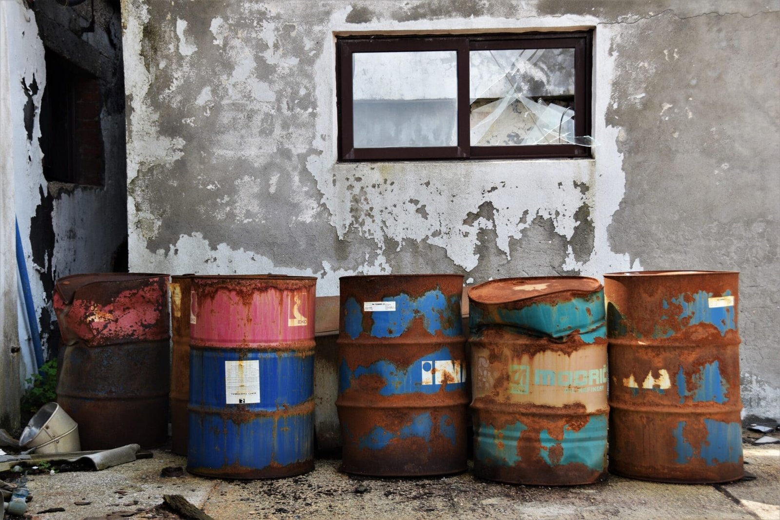 colourful, rusting barrels in front of a white wall with a broken window