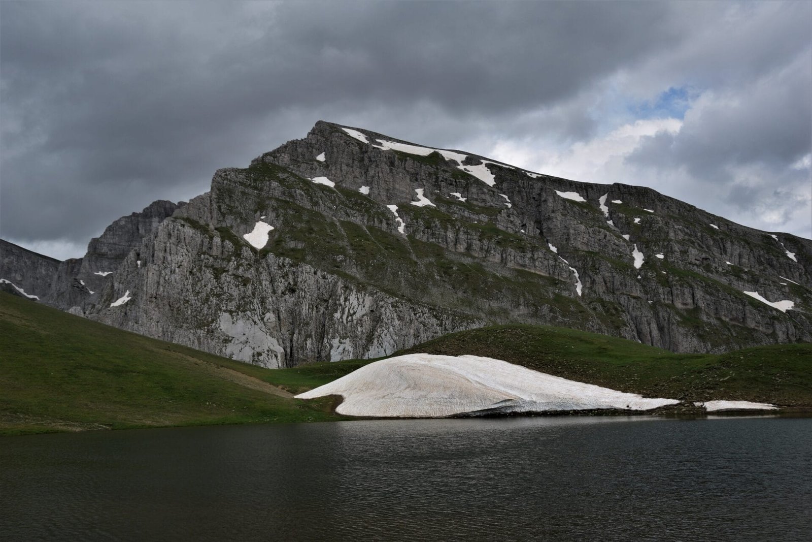 an imposing peak looms over an alpine mountain lake surrounded by green meadows and a left-over snowfield