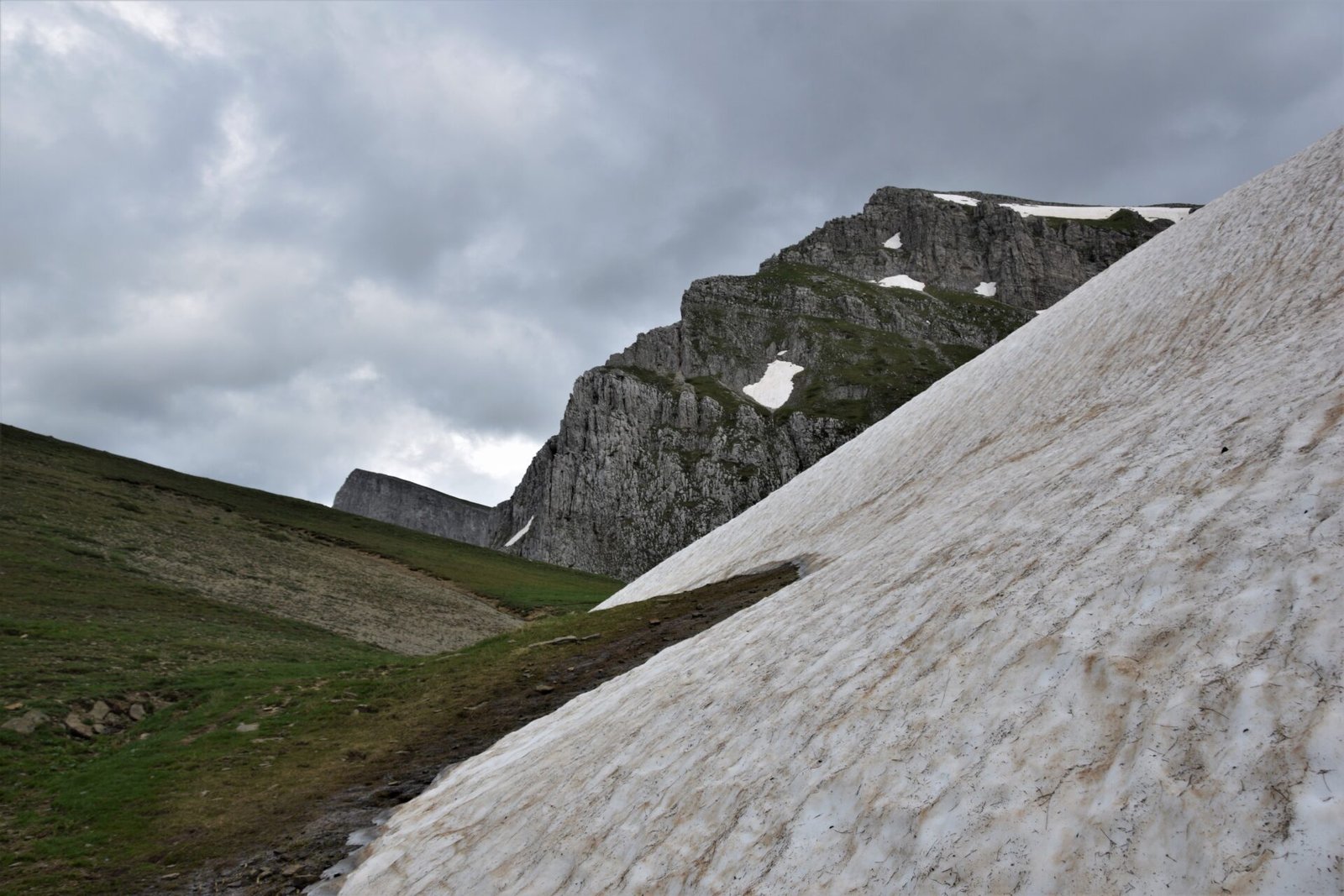 imposing peaks loom behind a green alpine meadow half covered in snow