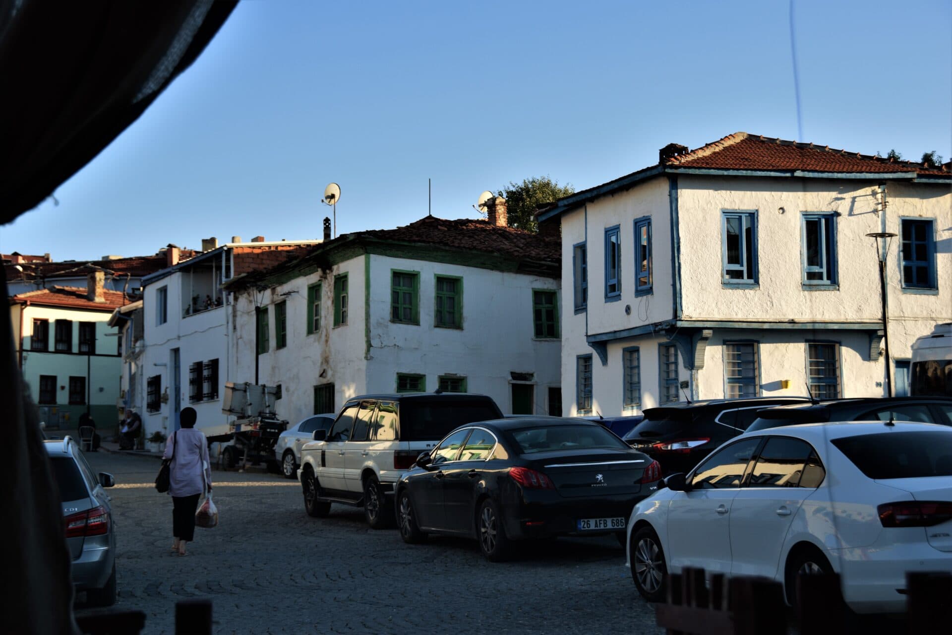 a woman walks between parking cars towards whitewashed buildings boasting blue and green coloured window and door frames