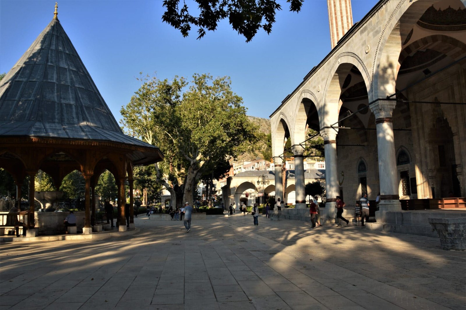 people walk across the open space between a mosque and ablution fountain covered by a wooden gazebo