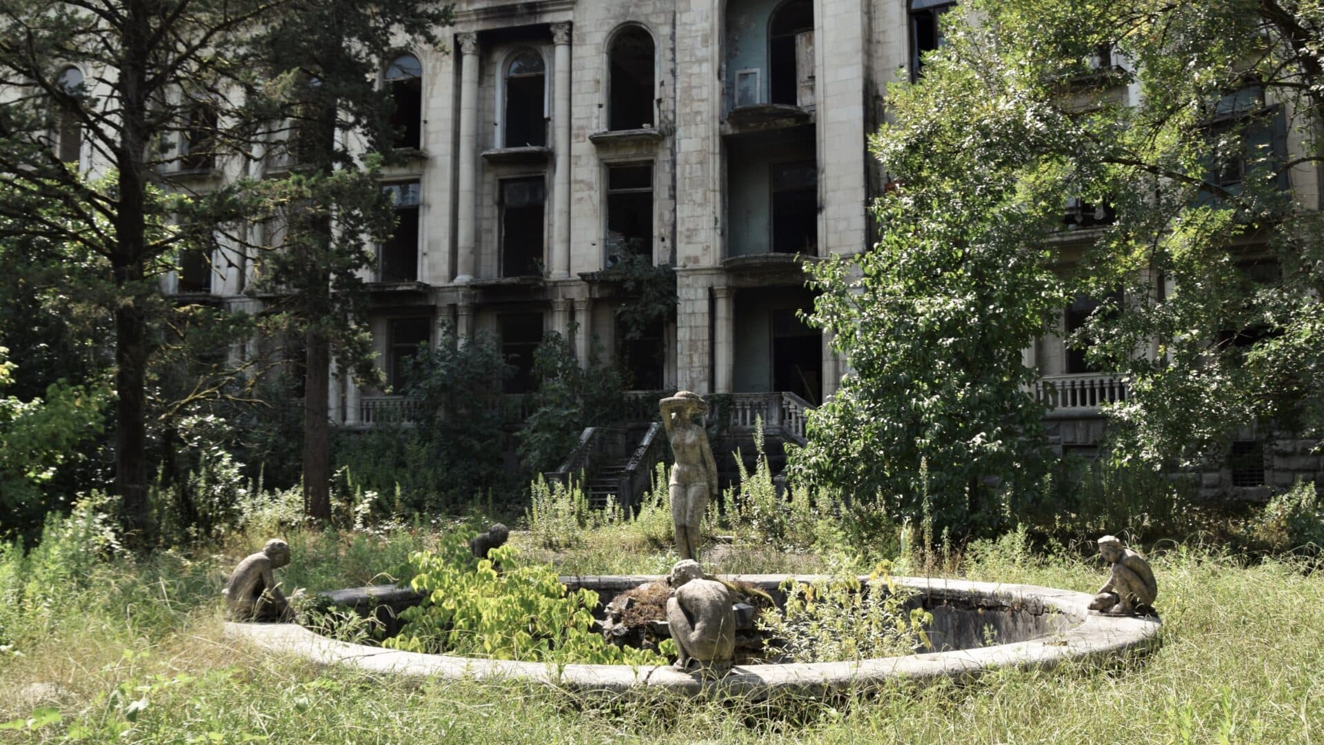 an overgrown fountain, boasting statues of four boys sitting on its edge and a girl in the middle, stands derelict in front of an abandoned Soviet sanatorium